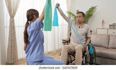 Asian Personal Resident Care Attendant Helping Retired Male Wheelchair User Practicing Physical Therapy With Resistance Band. Senior Fitness At Home Concept