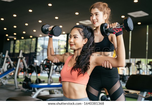 Asian personal trainer coaching a bodybuilding\
woman to perform the weightlifting exercise with dumbells in the\
fitness gym. Weightlifting and workout training coach business\
concept