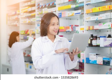 Asian Pharmacist Female Using Laptop Computer And Checklist Inventory Or Reading Information Medication In Pharmacy Or Drugstore. Medicine, Pharmaceutics, Health Care And People Concept 