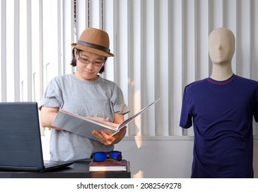 Asian Plus Size LGBT Female Designer In Gray T-shirt With Fedora Hat Is Looking Fashion Magazine With Laptop Computer On The Table In Home Office Room