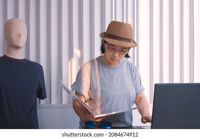 Asian Plus Size LGBT Female Designer In Gray T-shirt With Fedora Hat Using Laptop Computer While Looking At Fashion Magazine At Home Office In Evening Time