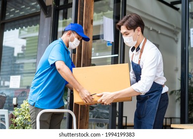 Asian Postman Delivery In Blue Uniform Send Food Ingredients Box Deliver To Restaurant Waiter In Coffeehouse. Attractive Barista Worker Wear Mask Due To Covid-19, Sign For Parcel And Carry Into Cafe.