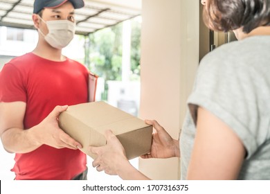 Asian Postman Or Deliveryman Carry Small Box Deliver To Young Woman Customer In Front Of Door At Home. Man Wearing Mask Prevent Covid Or Coranavirus Affection Outbreak. Social Distancing Work Concept.