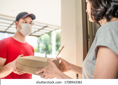 Asian Postman Or Deliveryman Carry Small Box Deliver To Young Woman Customer In Front Of Door At Home. Man Wearing Mask Prevent Covid Or Coranavirus Affection Outbreak. Social Distancing Work Concept.