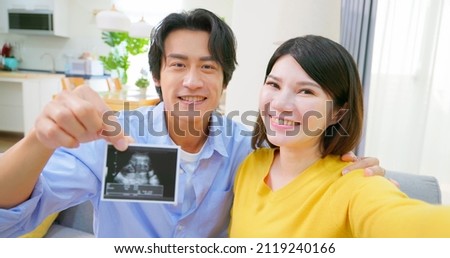 asian pregnant couple sitting on couch have video chat with friends through mobile phone together and showing their baby ultrasound photograph in living room at home