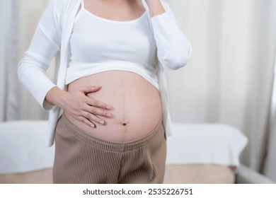 Asian pregnant woman adjusting headphones on baby bump in softly lit room. Calm expression showing connection with unborn child, enjoying soothing music during late pregnancy. - Powered by Shutterstock