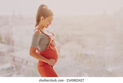 Asian pregnant woman happy touching her pregnancy bump for prenatal photoshoot wearing maternity clothes overalls. Beautiful young multiracial girl model