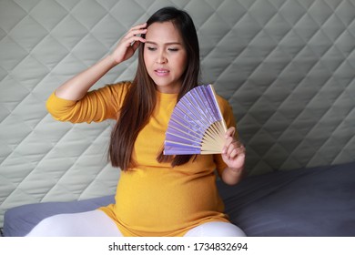 Asian Pregnant Woman Having Hot Flashes And Fanning Herself On Bed At Home. Pregnancy Suffering From Heat And Sweat.last Trimester Expecting.