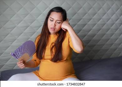 Asian Pregnant Woman Having Hot Flashes And Fanning Herself On Bed At Home. Pregnancy Suffering From Heat And Sweat.last Trimester Expecting.