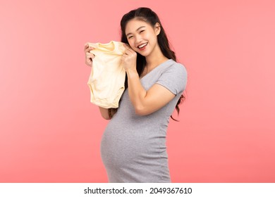 Asian Pregnant Woman Holding Baby Cloth For Baby Newborn Isolated On Pink Background. Pregnant Woman Packing Baby Stuff Ready For The Maternity Hospital. Pregnancy Proud Prepare For Newborn Concept