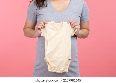 Asian Pregnant Woman Holding Baby Cloth For Baby Newborn Isolated On Pink Background. Pregnant Woman Packing Baby Stuff Ready For The Maternity Hospital. Pregnancy Proud Prepare For Newborn Concept