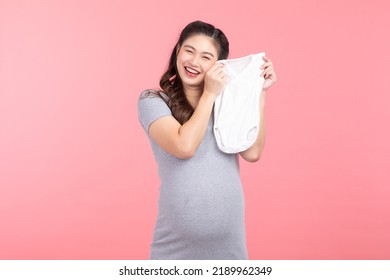 Asian Pregnant Woman Holding Baby Cloth For Baby Newborn Isolated On Pink Background. Pregnant Woman Packing Baby Stuff Ready For The Maternity Hospital. Pregnancy Proud Prepare For Newborn Concept