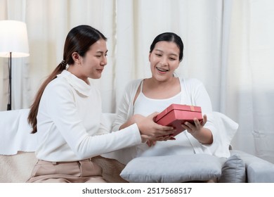 Asian pregnant woman receiving a red gift box from a friend. Smiling and expressing surprise. Warm and caring atmosphere, symbolizing friendship and support during pregnancy. Cozy home setting. - Powered by Shutterstock