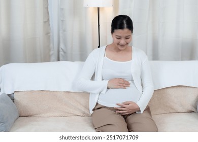 Asian pregnant woman sitting on sofa smiling while gently touching her belly, expressing love and care for unborn baby. Calm and peaceful, bright white decor enhancing, reflecting anticipation and joy - Powered by Shutterstock