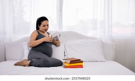 Asian pregnant woman sitting on a bed holding headphones, with a stack of colorful books nearby, promoting relaxation and prenatal care - Powered by Shutterstock