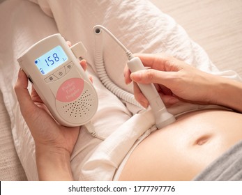 Asian Pregnant Woman Using Pocket Fetal Doppler To Monitor Baby Heart Beat On Sofa Couch At Home. Expectant Mother Happy And Smile When Examine Belly.