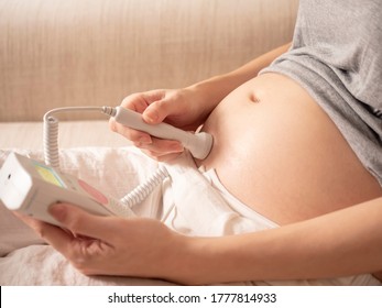 Asian Pregnant Woman Using Pocket Fetal Doppler To Monitor Baby Heart Beat On Sofa Couch At Home. Expectant Mother Happy And Smile When Examine Belly.