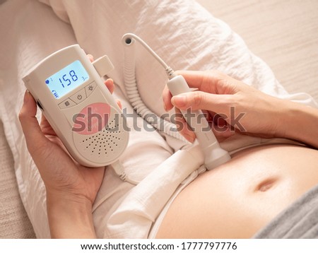 asian pregnant woman using pocket fetal doppler to monitor baby heart beat on sofa couch at home. expectant mother happy and smile when examine belly.
