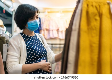 Asian Pregnant Woman Wear Face Mask Shopping In A Boutique, Young Female Looking And Choosing Baby Clothing In A Shop.