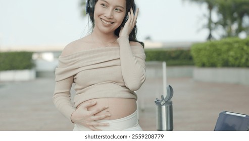 Asian pregnant woman wearing headphone listening to music smiling sitting outdoor. pregnancy mom take care, mental health, happy. single mother caresses belly make unborn baby feel good. Health care - Powered by Shutterstock