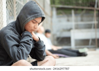 Asian pre-teen boy in a black hoodie sits with his knees pressed against a metal fence panel in a juvenile detention facility, awaiting further release, freedom and detention of people concept. - Powered by Shutterstock