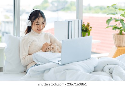 Asian pretty female teenager girl in turtleneck sweater wearing headphones listen to music from laptop on white clean sheet bed under blanket drinking coffee with cute domestic tabby short hair cat. - Shutterstock ID 2367163861