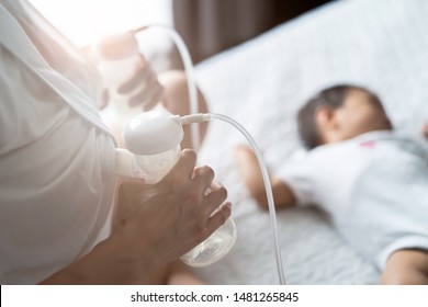 Asian pretty mother pumping breast milk on the bed in bedroom. The baby lying and sleeping on the bed beside her. Mother holding the milk and looking the child with love and smiling. Pump milk concept