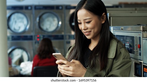 Asian pretty woman with long dark hair tapping and texting message on smartphone while standing in public laundromat.  - Powered by Shutterstock