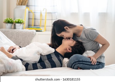 Asian pretty woman sitting on bed bending face down and kissing boyfriend lying on her leg, looking on eyes and smile feeling love very much. Romantic moment of couple lover in Valentine day concept.
