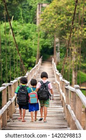 Asian Primary School Girls In Casual Clothes Walking Cross A Simple Suspension Footbridge At Dusk, Rural Scene Near Thailand And Myanmar Border. Education Concept.