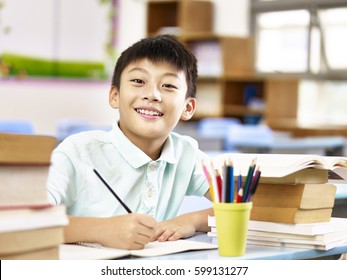 Asian Primary School Student Looking At Camera Smiling While Studying In Classroom.