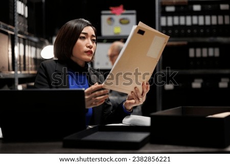 Asian private detective in agency file cabinet room reading criminal case file details. Investigator woman surrounded by criminology document folders on office repository cabinet shelves