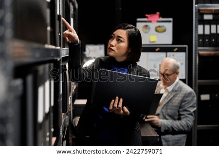 Asian private detective in criminal cases archive office looking for criminology file paperwork on cabinet shelves. Investigator woman surrounded by criminal case folders in agency repository room