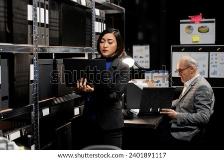 Asian private investigator in agency depository office room looking for criminal case file paperwork on cabinet shelves. Detective woman and elderly assistant coworker surrounded by criminology