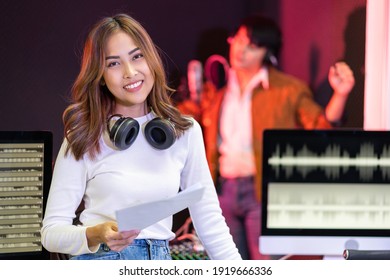 Asian Producer Woman In White Shirt Standing By Sound Mixing Console. Happy Female Music Composer Artist With A Man Singer Background