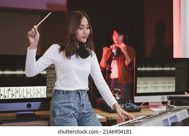 Asian Producer Woman In White Shirt Standing By Sound Mixing Console. Happy Female Music Composer Artist With A Man Singer Background  
