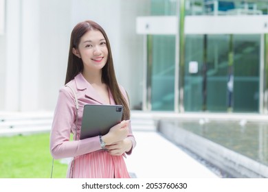 Asian Professional Business Female With Long Hair Is Smiling Outdoor In The Garden Front Of The Office While Hold At The Tablet In Her Hand To Go Out Meeting With Clients At The Office.