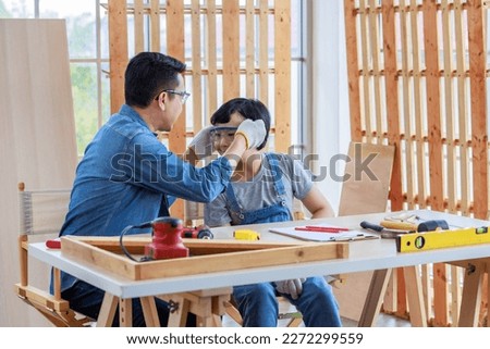 Asian professional carpenter engineer architect woodworker dad in jeans outfit with safety goggles gloves touching young boy son shoulder teaching mentering in housing home building construction site.