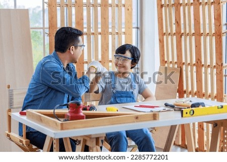 Asian professional carpenter engineer architect woodworker dad in jeans outfit with safety goggles gloves touching young boy son shoulder teaching mentering in housing home building construction site.