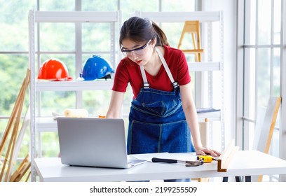 Asian Professional Female Engineer Architect Foreman Labor Worker Wears Safety Goggles And Jeans Apron Standing Smiling Using Laptop Computer Working On Wooden Table With Tools In Construction Site.