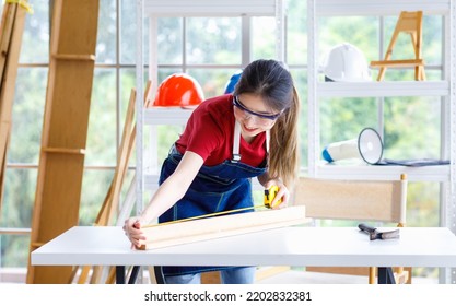 Asian Professional Female Engineer Architect Foreman Labor Worker Wears Safety Goggles Glasses Using Pencil And Square Angle Degree Ruler Measuring Wood Plank On Working Table With Laptop Computer.