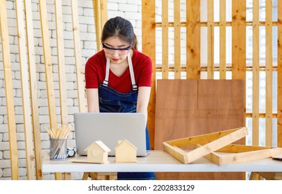 Asian Professional Female Engineer Architect Foreman Labor Worker Wears Safety Goggles And Jeans Apron Standing Smiling Using Laptop Computer Working On Wooden Table With Tools In Construction Site.