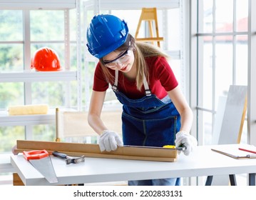 Asian Professional Female Engineer Architect Foreman Labor Worker Wears Safety Goggles Glasses Using Pencil And Square Angle Degree Ruler Measuring Wood Plank On Working Table With Laptop Computer.