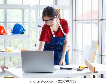 Asian Professional Female Engineer Architect Foreman Labor Worker Wears Safety Goggles And Jeans Apron Standing Smiling Using Laptop Computer Working On Wooden Table With Tools In Construction Site.