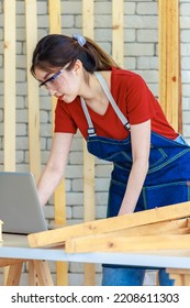 Asian Professional Female Engineer Architect Foreman Labor Worker Wears Safety Goggles And Jeans Apron Standing Smiling Using Laptop Computer Working On Wooden Table With Tools In Construction Site.