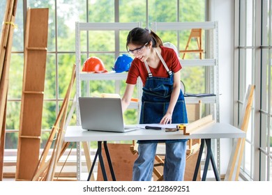 Asian Professional Female Engineer Architect Foreman Labor Worker Wears Safety Goggles And Jeans Apron Standing Smiling Using Laptop Computer Working On Wooden Table With Tools In Construction Site.