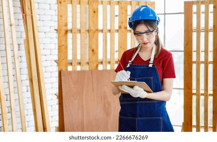 Asian professional female engineer architect foreman inspector wears safety goggles hard helmet jeans apron and gloves standing using pencil writing note on paper clipboard in construction site. - Powered by Shutterstock