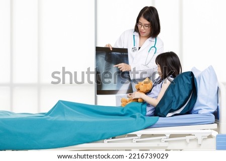 Asian professional friendly female doctor in white lab coat and stethoscope standing smiling showing lungs Xray film to young girl patient lay down on bed hugging teddy bear doll in hospital wardroom.