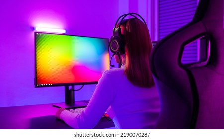 Asian Professional Gamer Playing Online Video Game On Desktop Computer PC Have Colorful Neon LED Lights Dark Room, Young Woman In Gaming Headphones Using Computer For Playing Game At Home, Back View