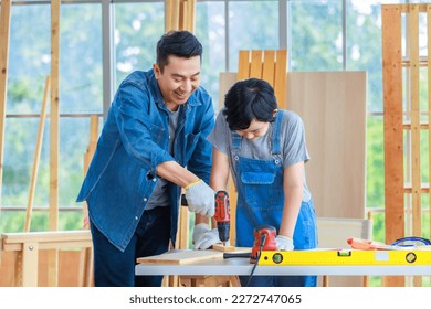 Asian professional male carpenter woodworker engineer dad in jeans outfit with safety gloves teaching young boy son using machine electric drill digging wooden stick in housing construction site.